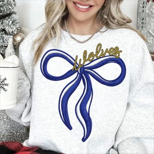 May include: A light gray sweatshirt featuring a large blue bow with white accents and the word "Wolves" in gold script. The bow is embroidered on the front of the sweatshirt.