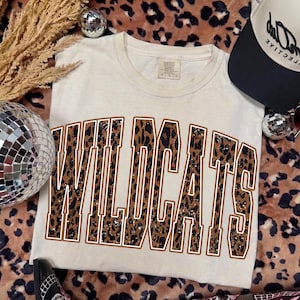 May include: A cream-colored short-sleeve t-shirt featuring the word WILDCATS printed in a large, arched collegiate font. The letters have a brown and black leopard print pattern with a thin white outline. The shirt has a crew neckline.