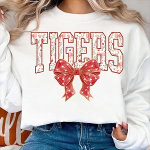 May include: White sweatshirt featuring the word "TIGERS" in a floral and heart-filled design. A large red bow with white hearts and gold accents is centered below the text. The sweatshirt is a casual, comfortable style.