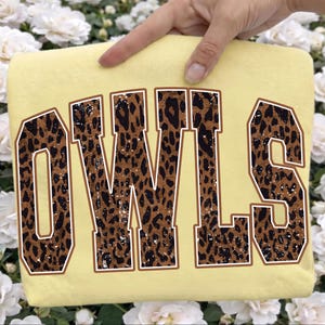 May include: A pale yellow t-shirt featuring the word OWLS in large, block-style varsity lettering. The letters have a brown and black leopard print pattern with a distressed, weathered texture and a thin white outline around each character.