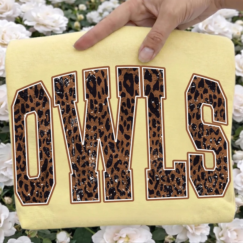 May include: A pale yellow t-shirt featuring the word OWLS in large, block-style varsity lettering. The letters have a brown and black leopard print pattern with a distressed, weathered texture and a thin white outline around each character.