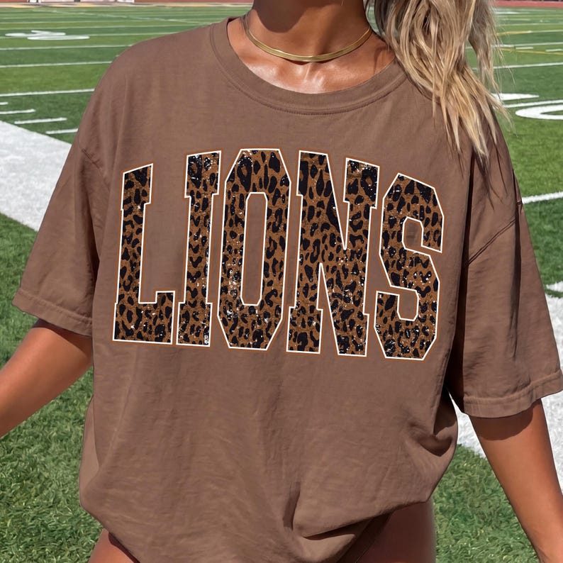 May include: A brown short-sleeved t-shirt featuring the word LIONS printed in large, block-style letters across the chest. The letters have a brown and black leopard print pattern with a thin white outline, creating a bold graphic design.