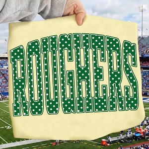 May include: A pale yellow t-shirt featuring the word ROUGHERS printed in large, bold, dark green block letters. The letters are decorated with a repeating pattern of small white polka dots, creating a distinct contrast against the fabric.