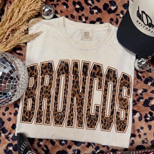 Puede incluir: Una camiseta de manga corta de color crema con la palabra BRONCOS en letras grandes de estilo bloque. Las letras tienen un estampado de leopardo marrón y negro con un borde blanco. La camiseta tiene cuello redondo y una etiqueta de marca.