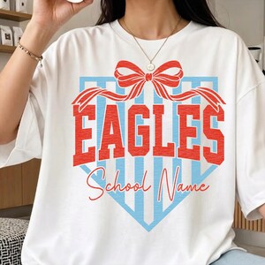以下が含まれることがあります： 赤色のブロック体で「EAGLES」の文字、赤いリボン、青と白のストライプの野球スタイルのグラフィックが特徴の白いTシャツ。「School Name」の文字は「EAGLES」の下に筆記体で書かれています。