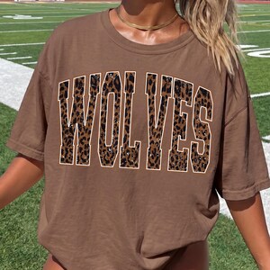 K&ouml;nnte beinhalten: Ein braunes T-Shirt mit kurzen &Auml;rmeln und dem Wort WOLVES, das in gro&szlig;en Blockbuchstaben aufgedruckt ist. Die Buchstaben haben ein braun-schwarzes Leopardenmuster mit einer d&uuml;nnen wei&szlig;en Umrandung, was ein auff&auml;lliges Design erzeugt.