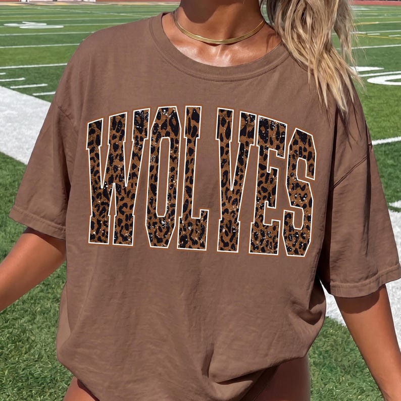 K&ouml;nnte beinhalten: Ein braunes T-Shirt mit kurzen &Auml;rmeln und dem Wort WOLVES, das in gro&szlig;en Blockbuchstaben aufgedruckt ist. Die Buchstaben haben ein braun-schwarzes Leopardenmuster mit einer d&uuml;nnen wei&szlig;en Umrandung, was ein auff&auml;lliges Design erzeugt.