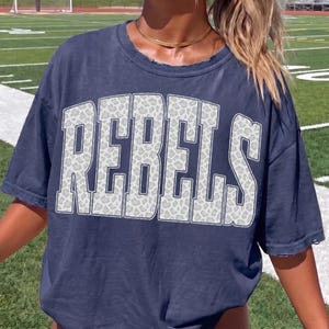 May include: Navy blue t-shirt with the word "REBELS" in large, white, leopard-print letters. The shirt has a distressed neckline and short sleeves. The background shows a green field.
