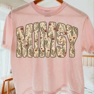 Op de afbeelding: Lichtroze t-shirt met het woord "MIMSY" in grote letters met bloemenpatroon. De letters zijn bruin omlijnd en gevuld met een beige achtergrond en een ontwerp van bloemen en bladeren. Het shirt hangt aan een houten hanger.