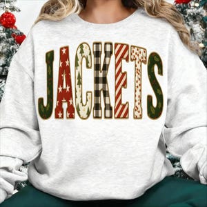 Peut inclure: Un sweat-shirt gris clair avec le mot "JACKETS" dans un design festif à motifs multiples. Les lettres présentent des étoiles rouges, du houx vert, du carreau noir et blanc et des rayures rouges et blanches, créant un thème de vacances.