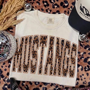 May include: A cream-coloured short-sleeve t-shirt featuring the word MUSTANGS in large, block-style letters. The letters have a brown and black leopard print pattern with a thin white outline. The shirt has a crew neckline and a tag at the collar.