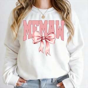 May include: White sweatshirt with the word "MEMAW" in pink gingham letters and a pink bow graphic. The sweatshirt is a crew neck style and has long sleeves. The design is centered on the front of the shirt.