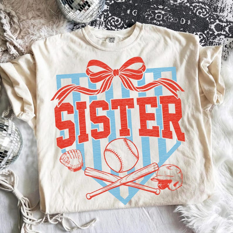 May include: Cream-colored t-shirt with a baseball-themed design. The word "SISTER" is in red block letters above a baseball, crossed bats, a glove, and a helmet. A red bow and blue stripes complete the design.
