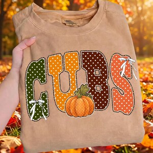 May include: A tan crewneck sweatshirt featuring the word CUBS in large, polka-dotted letters. The letters are green, orange, and brown with a leopard print outline. A small orange pumpkin sits in the center, and white bows accent the letters.