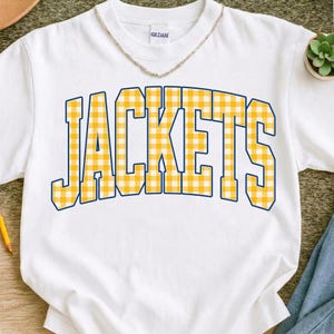 May include: White t-shirt with the word "JACKETS" in large, yellow and white gingham letters outlined in blue. The shirt has a crew neck and short sleeves. A gold chain necklace is visible at the neckline.