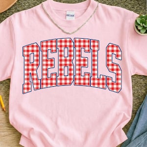 May include: Pink t-shirt with the word "REBELS" in large, bold letters across the front. The letters are in a red and white gingham pattern with a navy blue outline. The shirt has a crew neck and short sleeves.