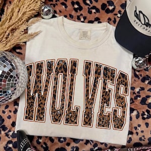 K&ouml;nnte beinhalten: Ein cremefarbenes Kurzarm-T-Shirt mit dem Wort WOLVES in gro&szlig;en Blockbuchstaben mit einem braun-schwarzen Leopardenmuster. Der Schriftzug ist mit einem d&uuml;nnen wei&szlig;en Rand versehen und das T-Shirt verf&uuml;gt &uuml;ber einen klassischen Rundhalsausschnitt.