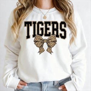 May include: White sweatshirt featuring the word "TIGERS" in black block letters with a leopard print fill. A leopard print bow is centered below the word. The sweatshirt is a crew neck style.
