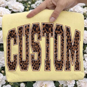 May include: A pale yellow t-shirt featuring the word CUSTOM in large, arched, varsity-style block letters. The letters have a brown and black leopard print pattern with a distressed texture and a thin white outline against the fabric.