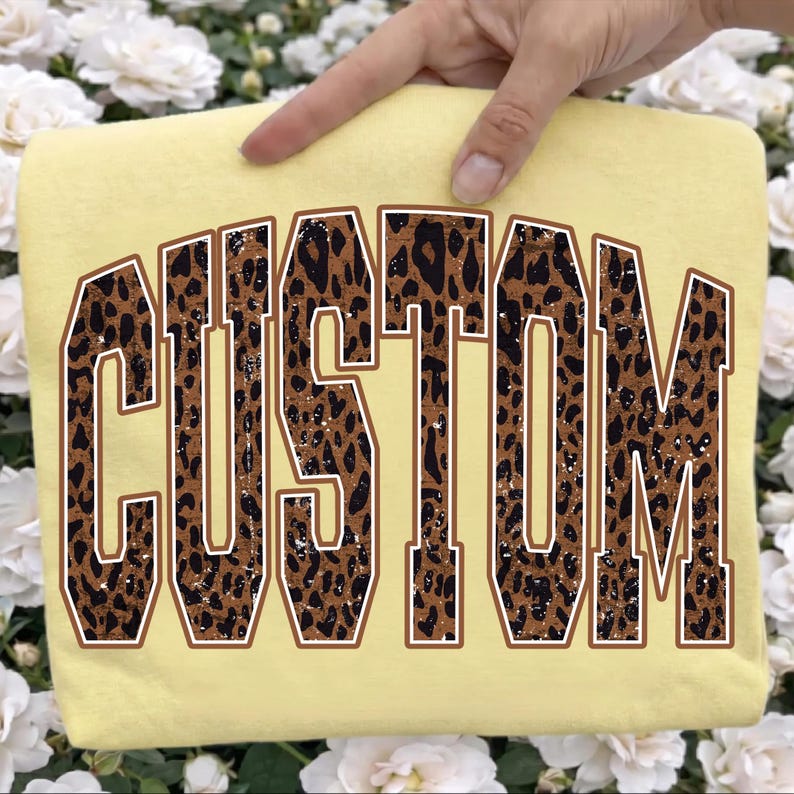 May include: A pale yellow t-shirt featuring the word CUSTOM in large, arched, varsity-style block letters. The letters have a brown and black leopard print pattern with a distressed texture and a thin white outline against the fabric.