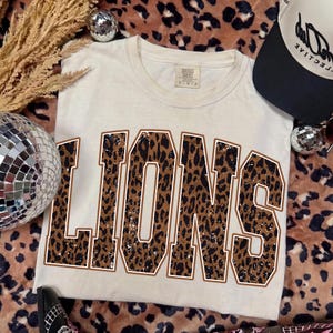 May include: A cream-colored short-sleeve t-shirt featuring the word LIONS printed in large block letters. The letters have a brown and black leopard print pattern with a white outline. The shirt has a crew neckline and a standard fit.