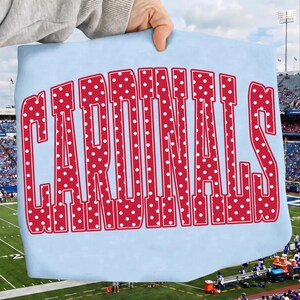 以下が含まれることがあります： 大きな赤い太字のブロック体で「CARDINALS」という文字がプリントされたライトブルーのTシャツ。赤い文字には小さな白い水玉模様が繰り返しあしらわれています。文字はわずかにアーチ状の大学風フォントで配置されています。