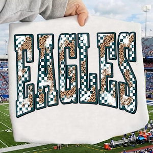 May include: A white t-shirt featuring the word EAGLES in large, block-style letters. The letters have a dark teal outline and are filled with a pattern that combines a brown and black leopard print with a white and dark teal checkered design.