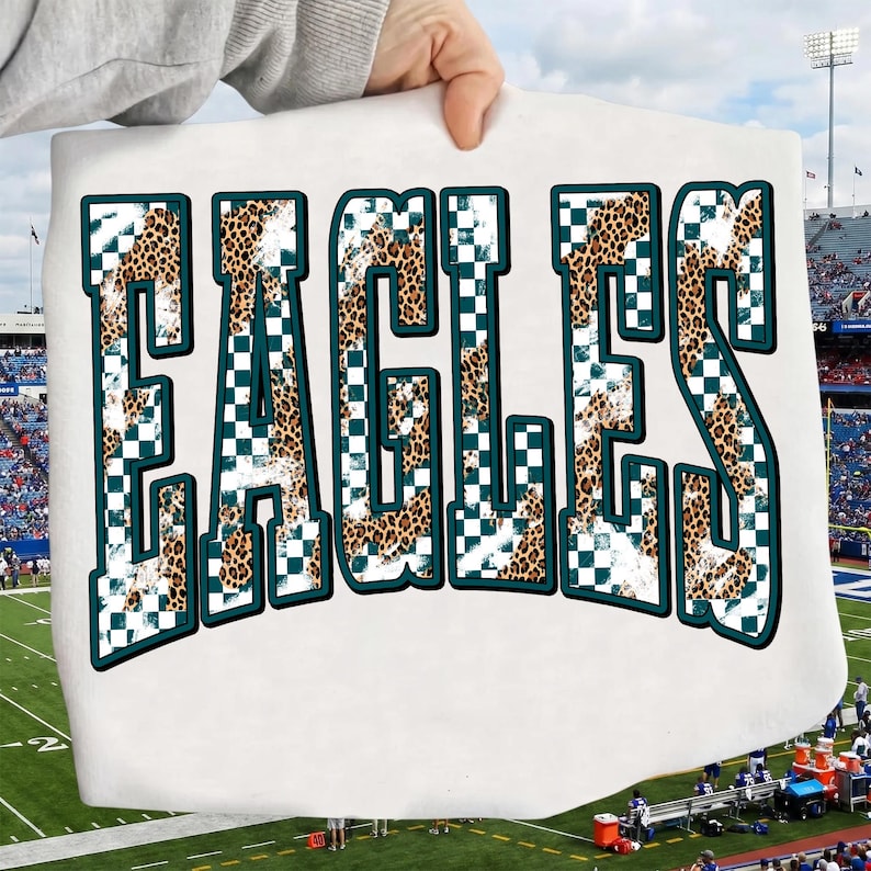 May include: A white t-shirt featuring the word EAGLES in large, block-style letters. The letters have a dark teal outline and are filled with a pattern that combines a brown and black leopard print with a white and dark teal checkered design.