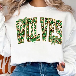 Peut inclure: Sweat-shirt blanc avec le mot "WOLVES" en grosses lettres. Les lettres sont remplies d'un motif léopard et de trèfles verts, soulignés d'or. Des nœuds verts et des trèfles complètent le design.