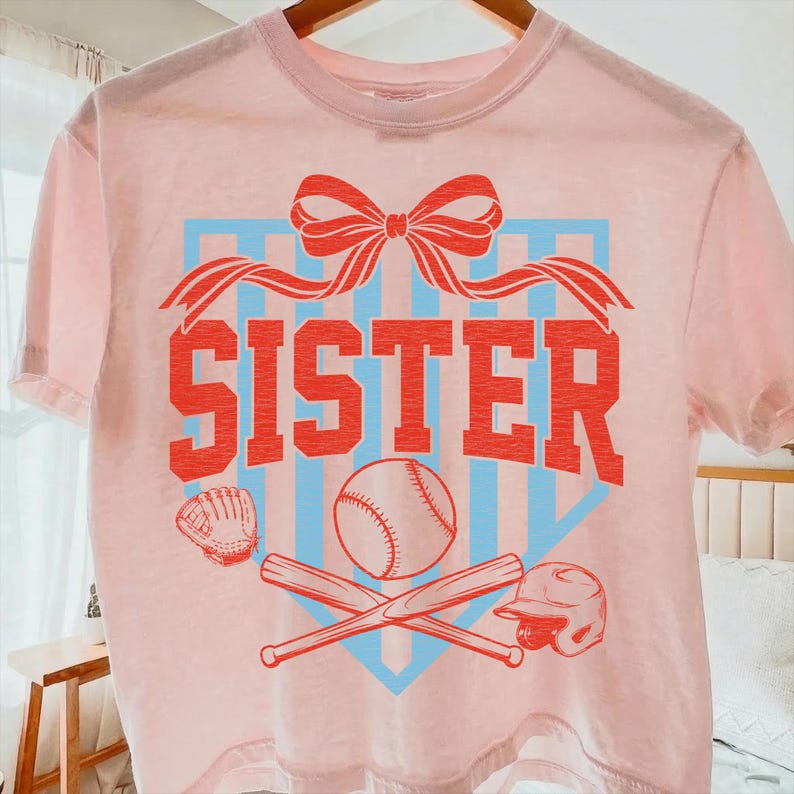 May include: A light pink t-shirt with a baseball-themed design. The word "SISTER" is printed in red, with a baseball, bats, glove, and helmet. A blue and red bow and ribbon design is at the top.