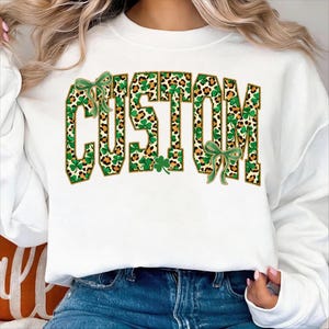 May include: White sweatshirt with the word "CUSTOM" in a green and leopard print design. The letters are outlined in gold, with green bows and shamrocks. The sweatshirt is a casual, everyday garment.