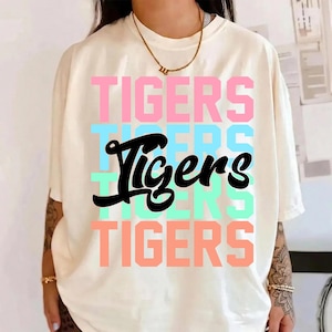 Peut inclure: T-shirt crème avec le mot "Tigers" superposé verticalement en rose, bleu, vert et pêche. Le mot "Tigers" est en gras, en noir au centre. Le t-shirt a des manches courtes.