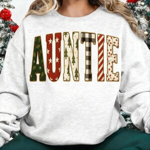 May include: A light grey sweatshirt featuring the word "AUNTIE" in a festive design. The letters are decorated with Christmas tree, star, and plaid patterns, outlined in gold. The sweatshirt is a casual, comfortable style, perfect for the holiday season.