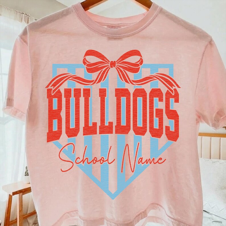 以下が含まれることがあります： 「BULLDOGS」の文字が赤いブロック体でプリントされた、薄いピンク色のTシャツ。赤いリボンと、縦線が入った青いひし形が文字の上と下に配置されています。「School Name」の文字は筆記体です。