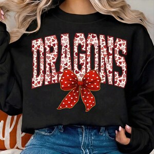 May include: Black sweatshirt with the word "DRAGONS" in a white font filled with red and pink hearts. A large red bow with white hearts is centered below the text. The sweatshirt is a casual style.