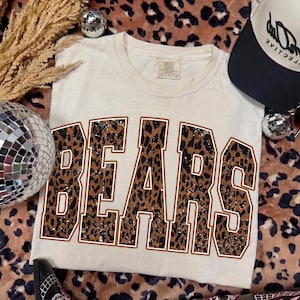 May include: A cream-colored short-sleeve t-shirt featuring the word BEARS in large, block-style letters. The letters have a brown and black leopard print pattern with a white speckled texture and a thin brown outline around each character.