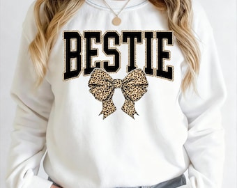 Op de afbeelding: Witte sweatshirt met het woord "BESTIE" in zwarte blokletters met een luipaardprint rand. Een luipaardprint strik is gecentreerd onder het woord. De sweatshirt is een crew neck stijl.