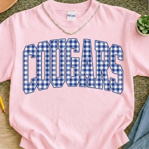 May include: Pink t-shirt with the word "COUGARS" in large, blue and white gingham letters across the front. The shirt has a crew neck and short sleeves. The brand "GILDAN" is printed on the tag.