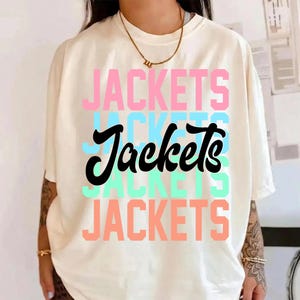 Può includere: T-shirt color crema con la scritta "JACKETS" in un design a strati. La parola è in rosa, blu, nero, verde menta e pesca. La maglietta ha maniche corte e girocollo.