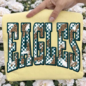 May include: A pale yellow t-shirt featuring the word EAGLES in large, block-style letters. The letters have a dark teal outline and are filled with a pattern of white and dark teal checkerboard squares mixed with a brown and black leopard print.