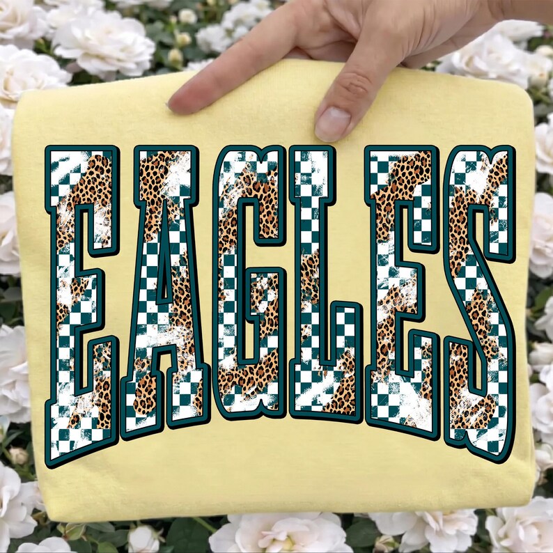 May include: A pale yellow t-shirt featuring the word EAGLES in large, block-style letters. The letters have a dark teal outline and are filled with a pattern of white and dark teal checkerboard squares mixed with a brown and black leopard print.