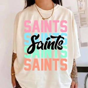 以下が含まれることがあります： ピンク、青、緑、オレンジ色の「SAINTS」の文字が縦に積み重ねられたクリーム色のTシャツ。「Saints」の文字は黒の筆記体です。Tシャツは半袖です。
