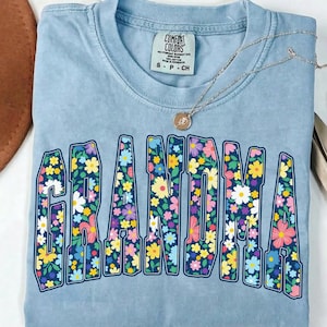 May include: A light blue Comfort Colors t-shirt with the word "GRANDMA" in large, floral-filled letters. A silver necklace with a pendant rests on the shirt. The shirt is folded neatly.