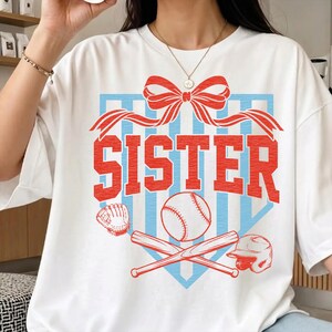 May include: White t-shirt with a baseball-themed graphic. The design features the word "SISTER" in red, a baseball, bats, a glove, and a helmet, all set against a blue and red background. A red bow is at the top.