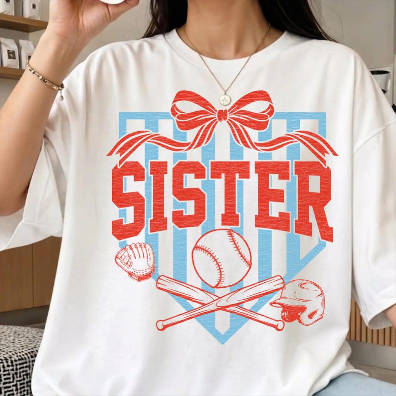 May include: White t-shirt with a baseball-themed graphic. The design features the word "SISTER" in red, a baseball, bats, a glove, and a helmet, all set against a blue and red background. A red bow is at the top.
