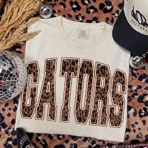 Könnte beinhalten: Ein cremefarbenes Kurzarm-T-Shirt mit dem Wort GATORS in großen Blockbuchstaben. Die Buchstaben haben ein braun-schwarzes Leopardenmuster mit weißer Umrandung. Das T-Shirt hat einen Rundhalsausschnitt und ein Etikett am Kragen.