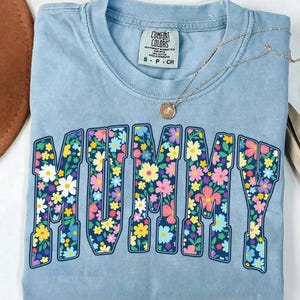 May include: A light blue t-shirt with the word "MUMMY" in large, floral-filled letters. The flowers are in shades of pink, yellow, blue, and white. A silver necklace with a pendant rests on the shirt. The shirt has a Comfort Colors tag.