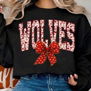 May include: Black sweatshirt with the word "WOLVES" in white letters filled with red and pink hearts. A large red bow with white hearts is centered below the word. The sweatshirt is worn with blue jeans.