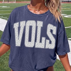 May include: Navy blue t-shirt with the word "VOLS" in large, white, block letters. The letters have a gray leopard print pattern. The shirt has a distressed neckline and short sleeves.