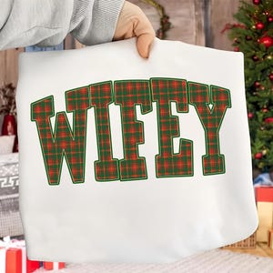 May include: A white tote bag with the word "WIFEY" in large, green-outlined letters filled with a red and green plaid pattern. The bag is held up by a person, with a Christmas tree visible in the background.